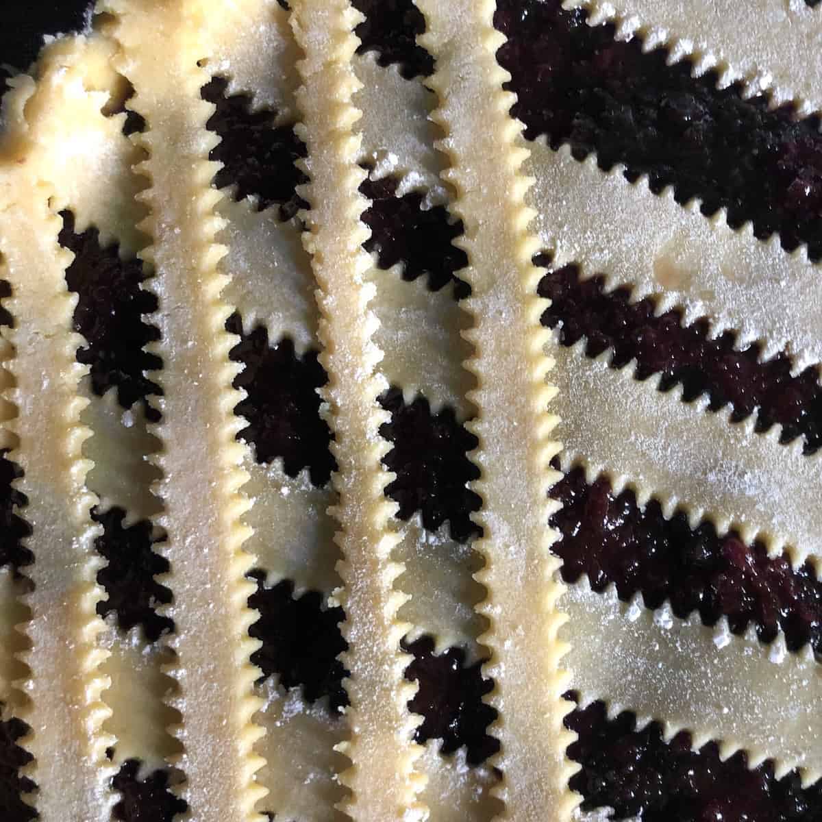 Closeup of adding reginelle lattice strips to crostata using easy trick to create lattice look without weaving or interlinking strands, simply laying pasta frolla strips in one direction then overlapping in opposite direction for authentic Italian jam tart appearance.