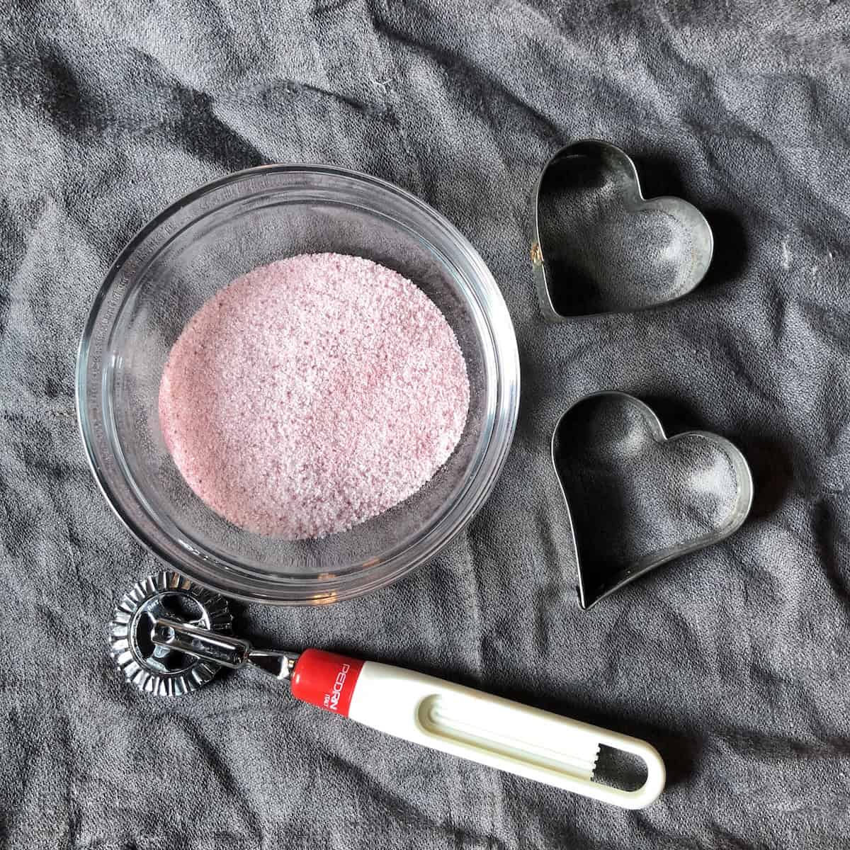 Homemade all-natural pink cherry sugar in a bowl made from freeze-dried cherries, displayed next to heart-shaped cookie cutters and pasta bike wheel with zigzag edges, tools for decorating Italian crostata di marmellata.