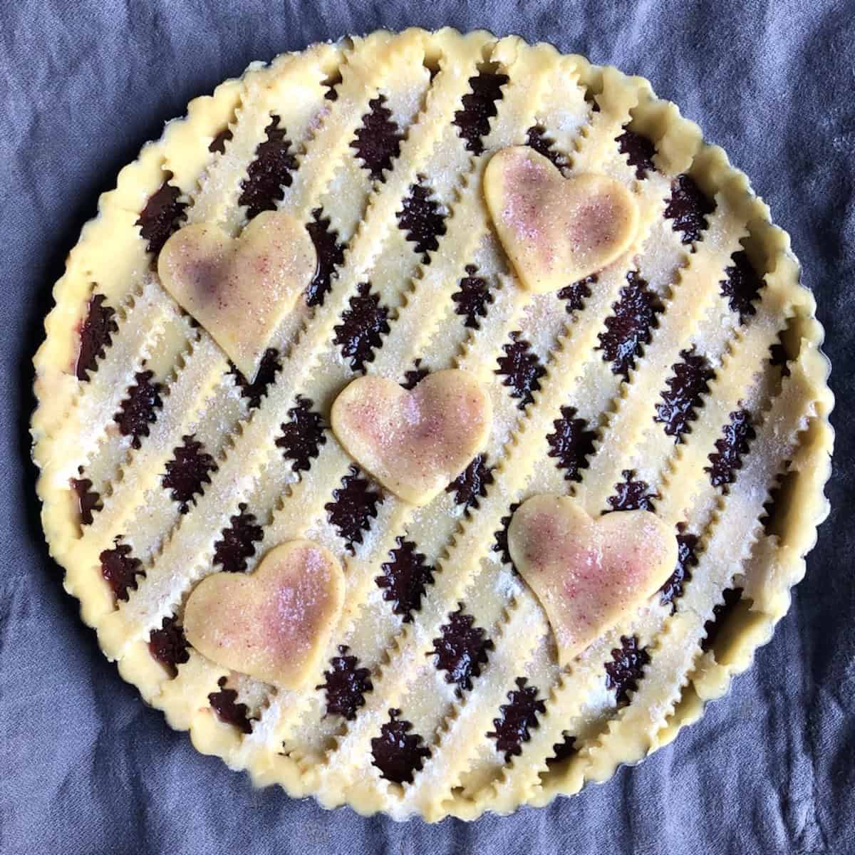 Unbaked, fully assembled homemade Italian crostata with reginelle zigzag lattice top, decorative heart cutouts, egg wash brushed on, and sprinkled with homemade cherry sugar, showing how to make authentic crostata di marmellata.