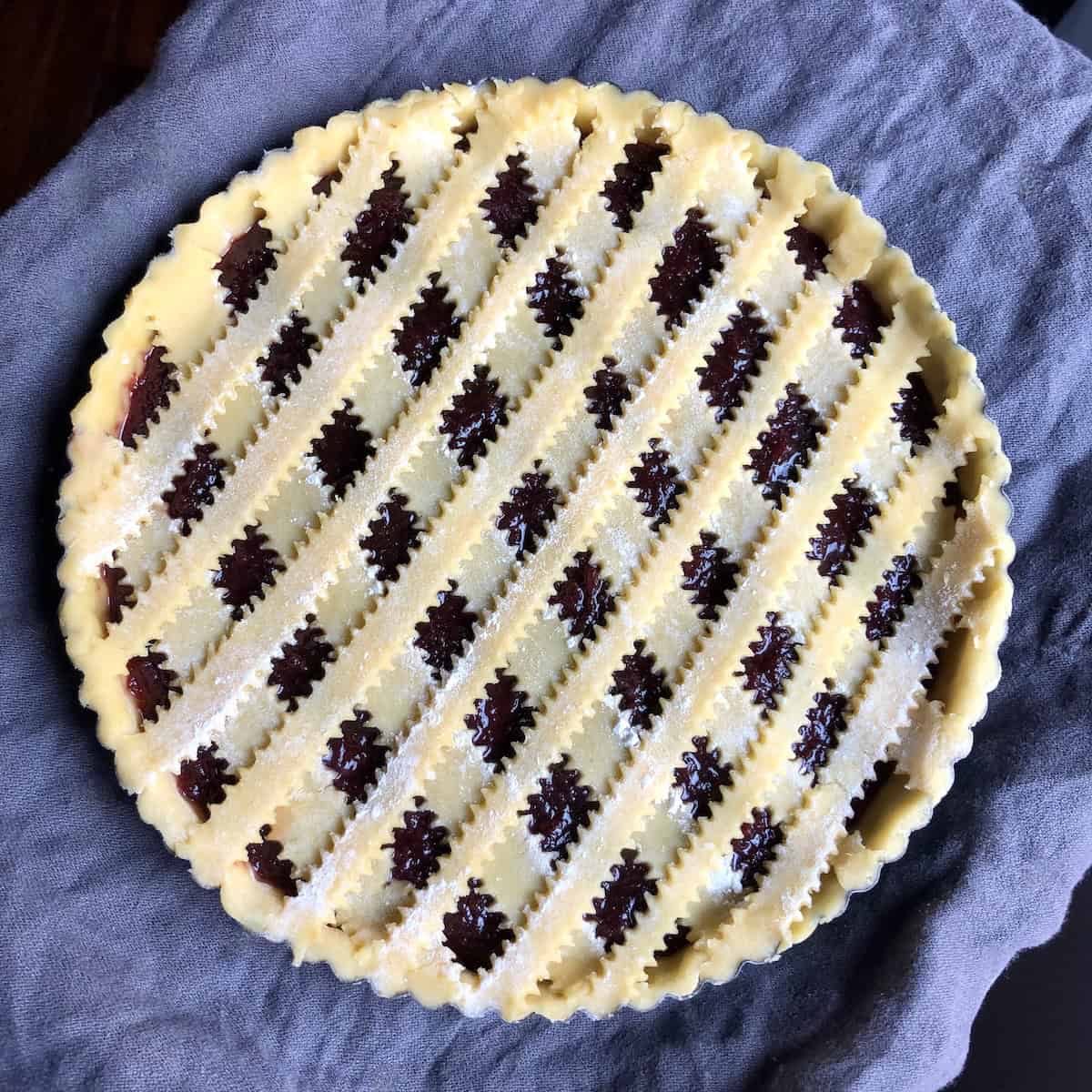 Homemade Italian crostata before baking with traditional reginelle zigzag-edged lattice crust top arranged in diamond pattern over jam filling, assembled pasta frolla dough ready for cutout decorations and egg wash, showing authentic Italian jam tart technique.