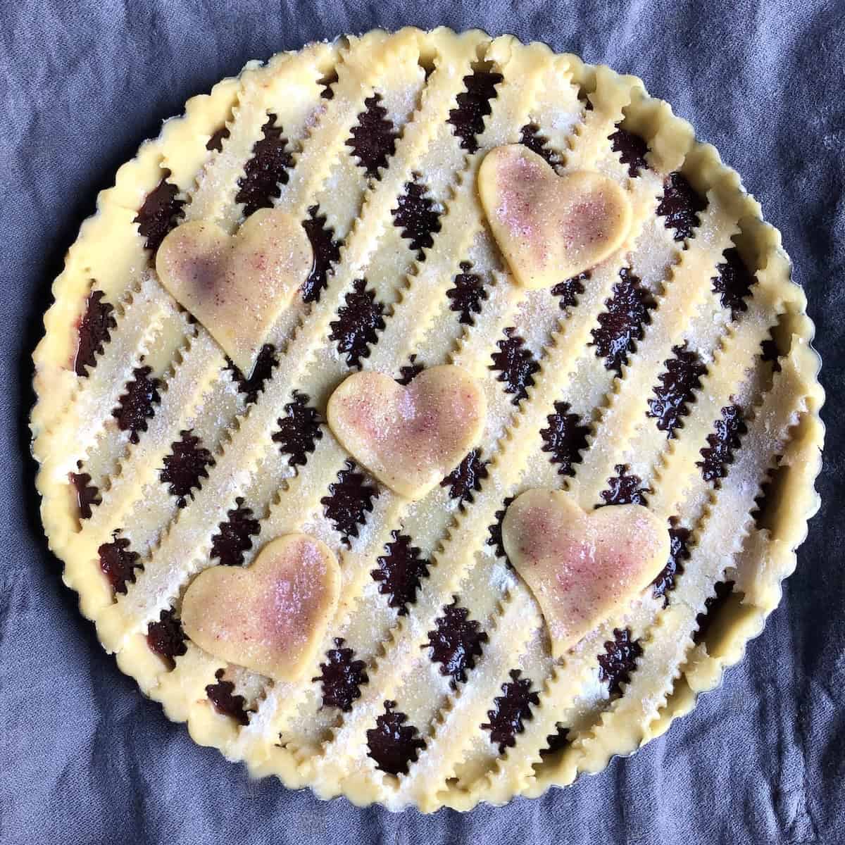 Fully assembled Italian crostata with reginelle zigzag lattice top, decorative pasta frolla heart cutouts, egg wash, and homemade pink cherry sugar sprinkled on top, ready to bake authentic crostata di marmellata.