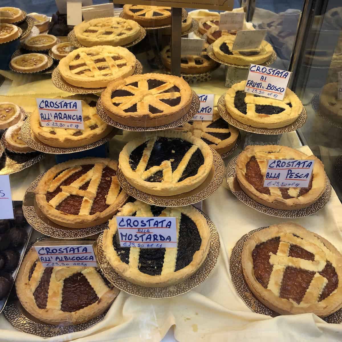 Italian crostata display at Paolo Atti & Figli pasticceria in Bologna showing authentic crostata di marmellata varieties including crostata all'albicocca (apricot crostata), crostata alla pesca (peach crostata), crostata ai frutti di bosco (wild berry crostata), crostata all'arancia (orange crostata), crostata alla mostarda, and chocolate crostata with golden lattice-topped pasta frolla crusts.