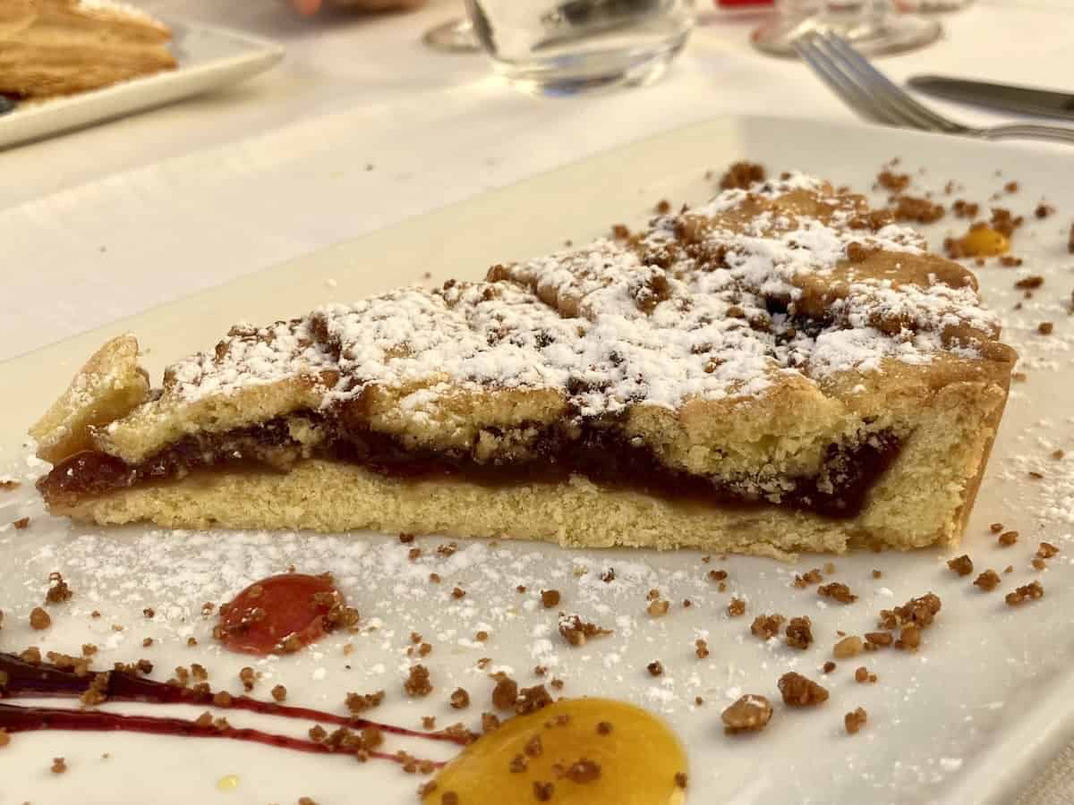 Italian strawberry crostata (crostata alla fragola) from a restaurant in the Colli Berici hills of Veneto, elegantly plated with strawberry coulis and pastry cream dots as garnish, featuring golden pasta frolla crust and vibrant strawberry jam filling.