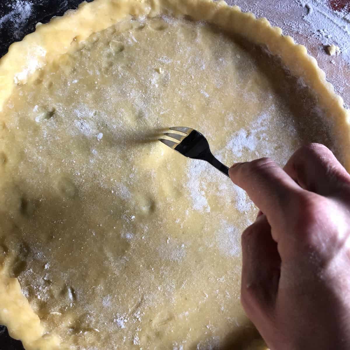 Using a fork to prick holes all over the bottom of pasta frolla crust to prevent air bubbles and puffing during baking, essential step for Italian crostata di marmellata jam tart.