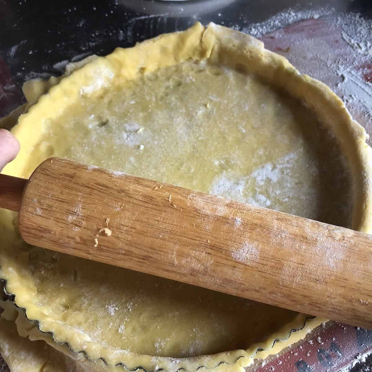 Pasta frolla dough pressed into tart pan with rolling pin rolled across top to cut off excess dough around edges, showing easy technique for trimming Italian crostata crust cleanly.