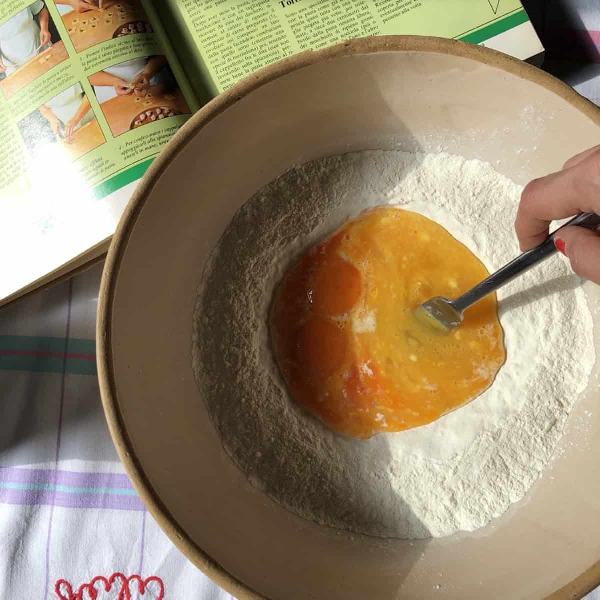 Using a fork to work eggs into a well of semolina and 00 flour in a vintage bread bowl with an Italian pasta cookbook in the background, semolina and 00 flour pasta recipe mixing step.