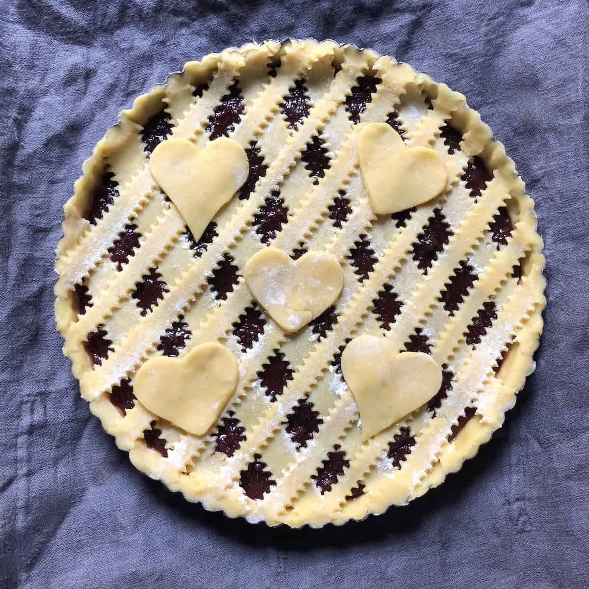 Fully assembled Italian crostata with perfect diamond lattice top pattern and decorative heart cutouts before egg wash and sugar are added, showing beautiful reginelle zigzag-edged pasta frolla strips ready for final touches before baking.