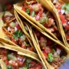 closeup of 5 corn tortilla steak tacos in a round cake pan held up by each other and topped with salsa, jalapeños, and cilantro