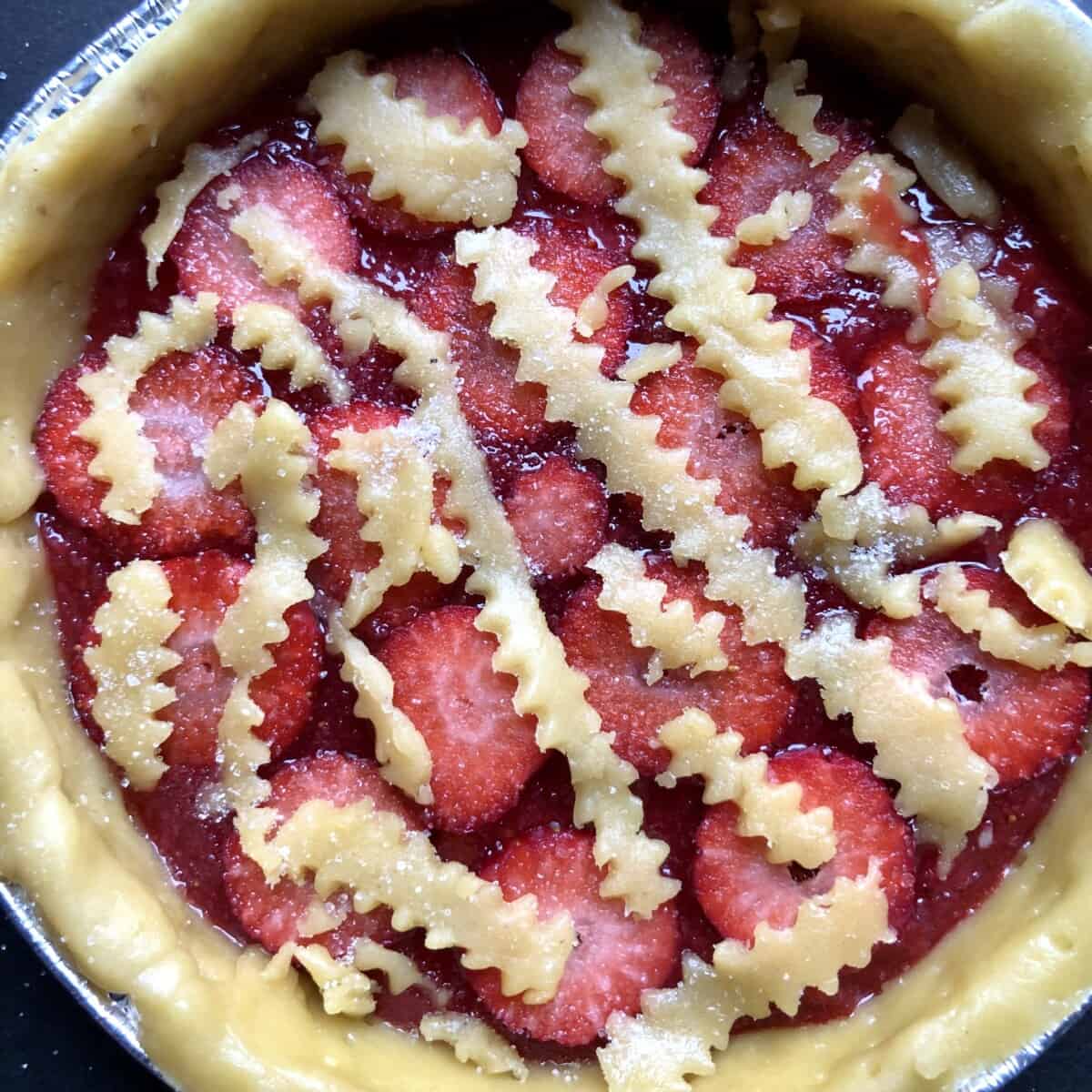 assembled and unbaked strawberry crostata with homemade strawberry jam and fresh sliced strawberries resting under leftover pieces of zigzagged edged lattice top