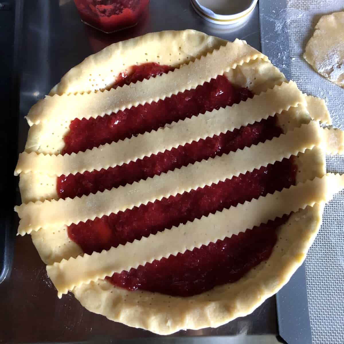 Adding the lattice top pasta frolla dough strips to the top of the strawberry crostata in a diagonal pattern to create traditional Italian diamond pattern.