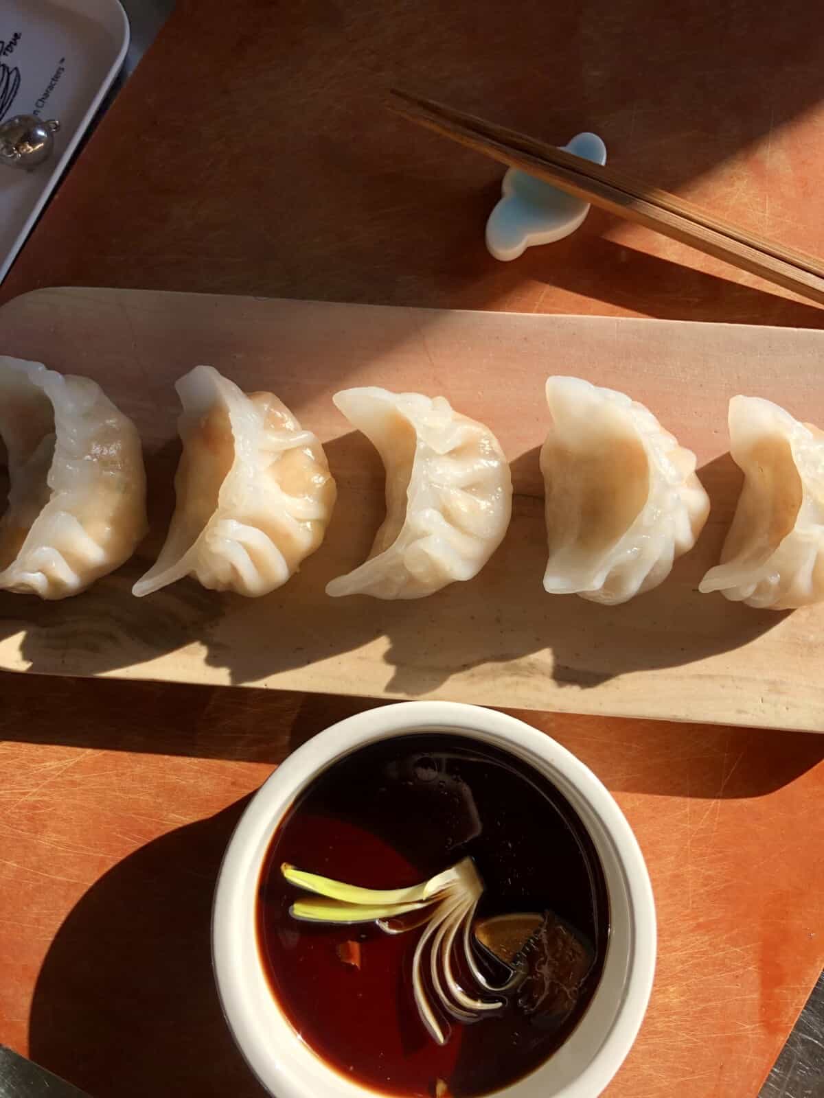 a pink marble platter with 5 har goa dumplings lined up on it and a side of dumpling dipping sauce with a flowered scallion and sliced ginger to flavor it