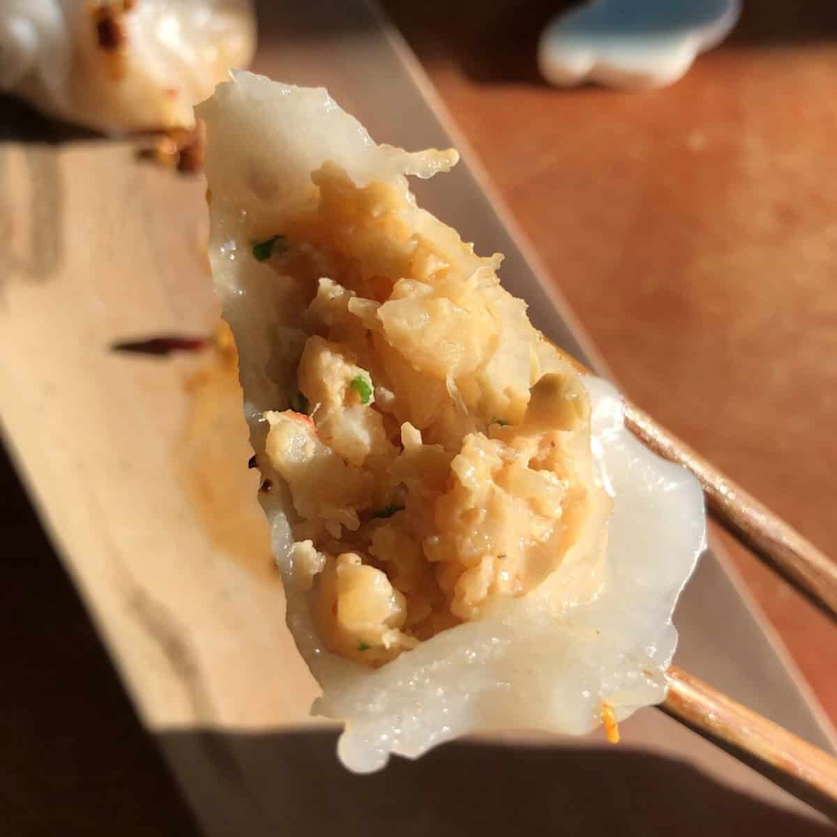 A bitten homemade har gow held in chopsticks showing the cooked shrimp filling inside the translucent wrapper.
