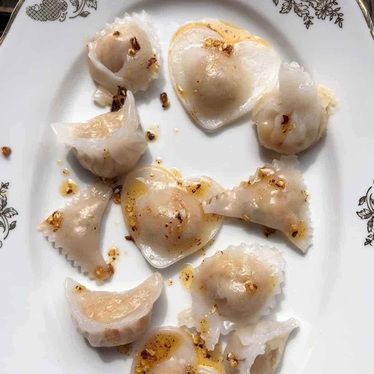 Eleven steamed har gow in various shapes including no-pleat triangle, heart, wonton, traditional pleated, and moneybag dumplings drizzled with Sichuan chili oil on a serving platter.
