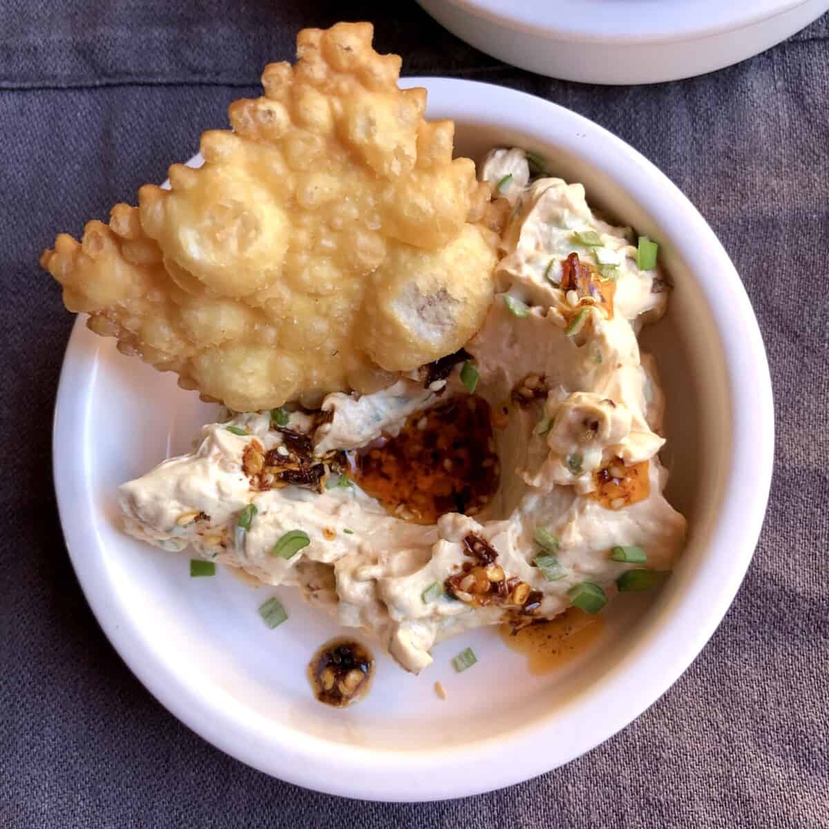 A small shallow ramekin dish with a dollop of chili oil cream cheese dip with a pool of extra chili oil drizzled in the middle well and a large homemade fried wonton chip tucked into the side.