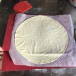 super thin rolled out 00 pizza dough that's been placed on a large piece of parchment paper with 2 flimsy plastic placemat/cutting boards undertneath for suppor