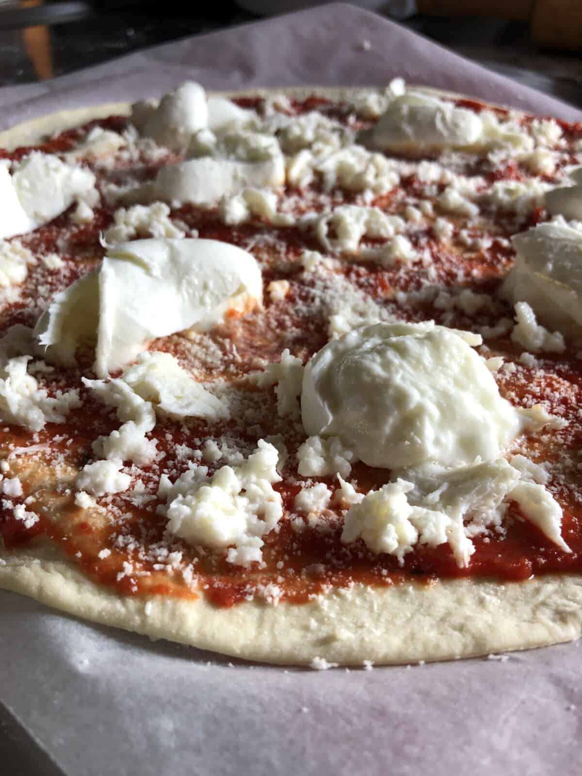 A closeup of a homemade pizza fully assembled with pizza sauce, freshly grated fior di latte (cow's milk mozzarella), and torn chunks of mozzarella di bufula (or buffalo mozzarella torn chunks on a very thin 00 flour pizza dough