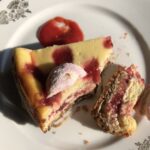 top down view of a plated piece of cheesecake speckled with strawberry dots o top, a homemade peep, and a piece on its side revealing the strawberry and strawberry cheesecake layers with Biscoff crust