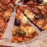 A sliced super crispy thin-crust pizza made using italian 00 flour and topped with spicy ventricina (similar to pepperoni), large cured black and green olives with buffalo mozzarella and whole milk mozzarella.
