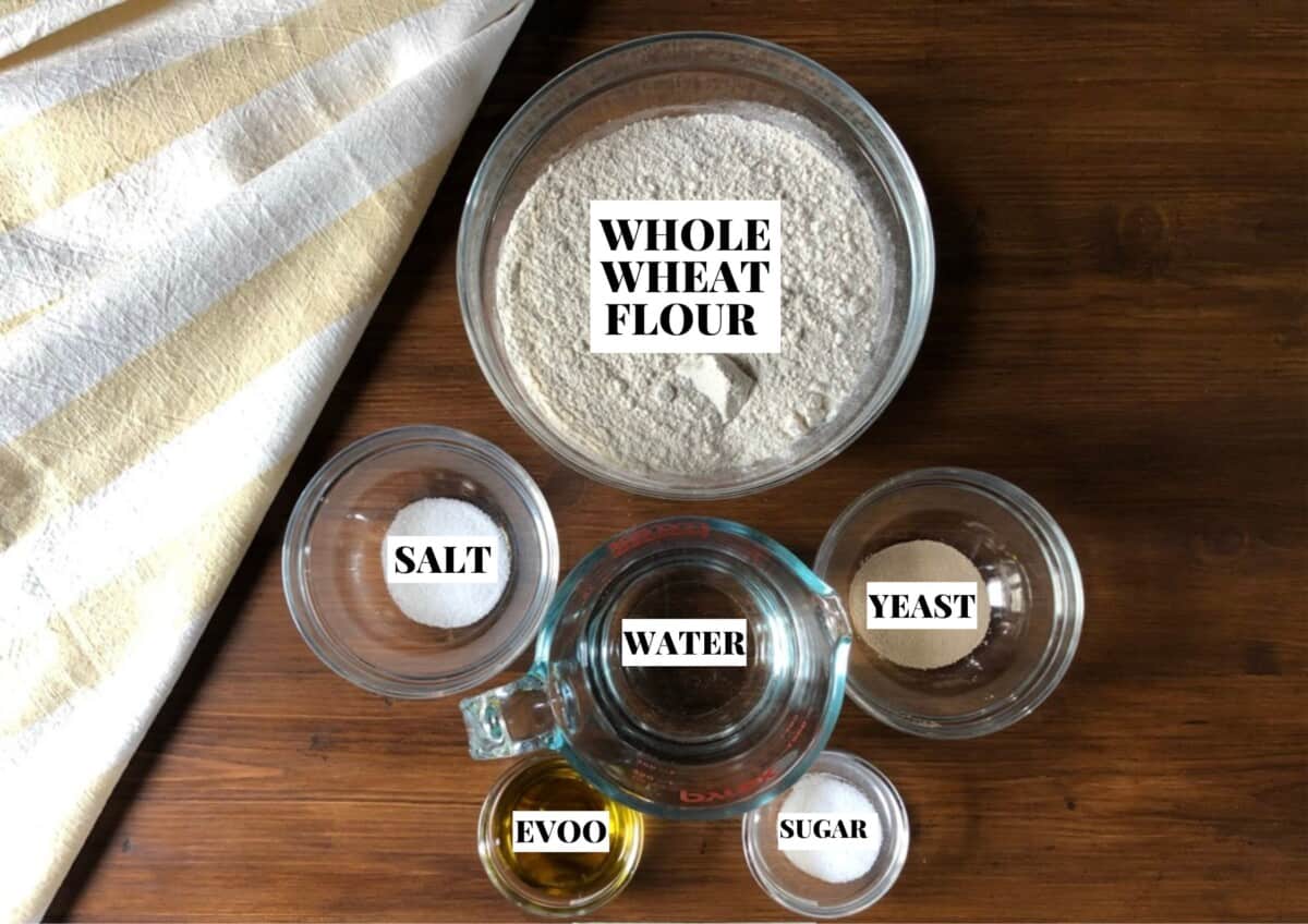 Whole wheat Pizza Dough ingredients on a table: whole wheat flour, yeast, sugar, extra virgin olive oil, salt, and water.