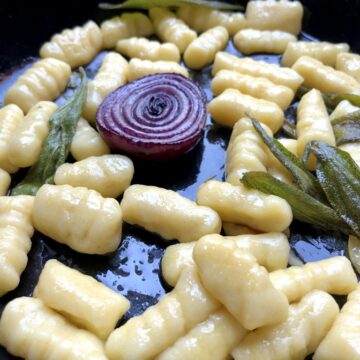 Homemade gnocchi in a cast iron skillet with half of a caramelized red onion and crispy sage leaves with extra virgin olive oil.