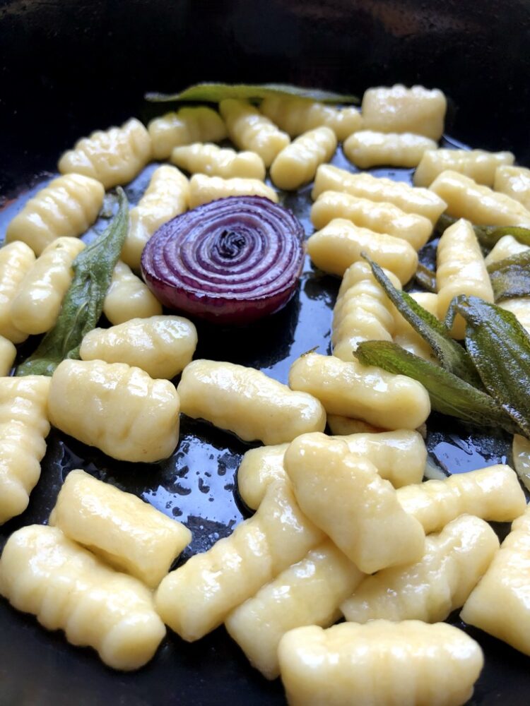 Homemade gnocchi in a cast iron skillet with half of a caramelized red onion and crispy sage leaves with extra virgin olive oil.