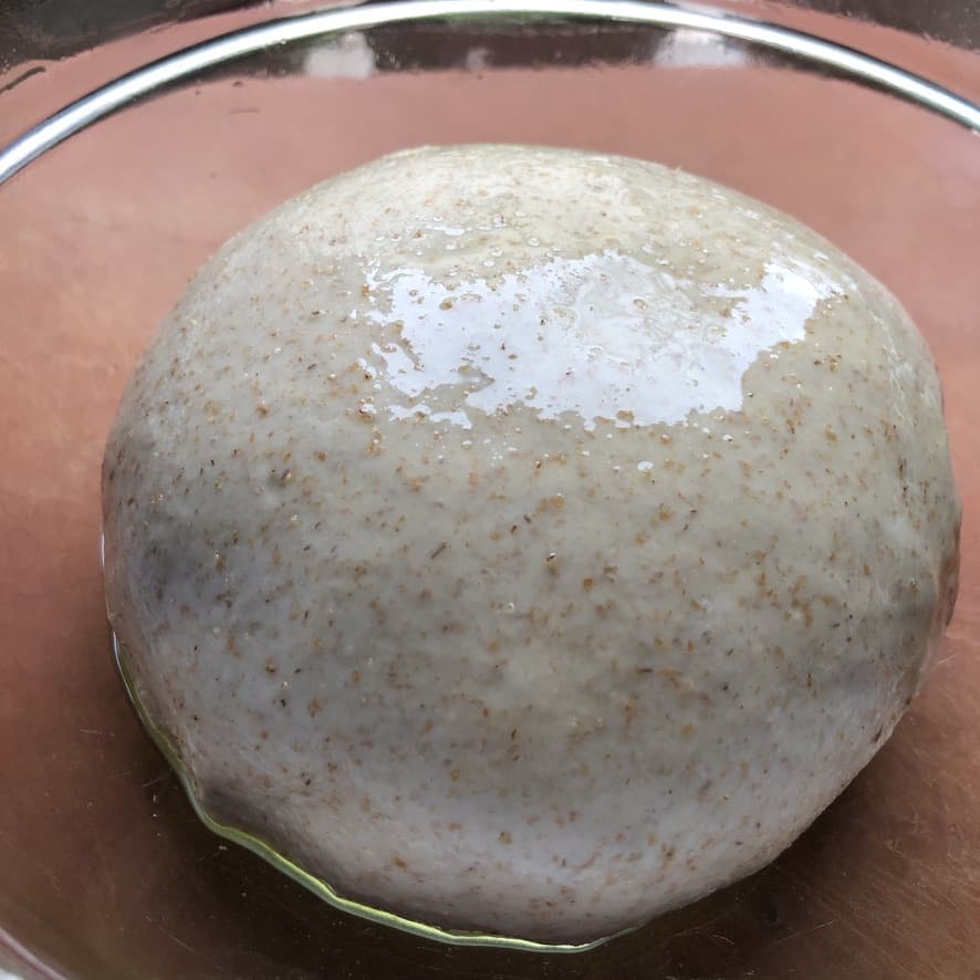 lightly oiled whole wheat pizza dough round in a clear glass bowl ready to be covered and left to rise for 90 minutes.