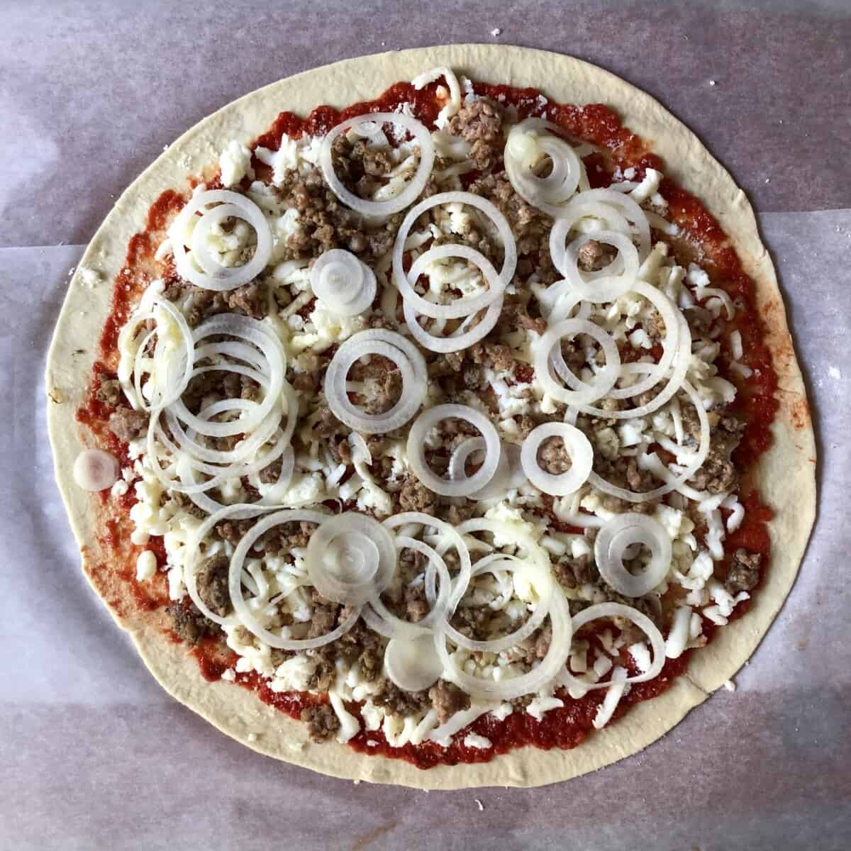 Fully assembled unbaked thin-crust pizza with sausage and onion toppings on bread flour dough.
