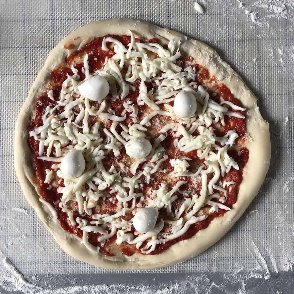Shredded whole milk mozzarella and baby buffalo mozzarella balls topping a bread flour pizza dough with homemade pizza sauce, how to make pizza at home with fresh cheese on a handmade pizza crust.
