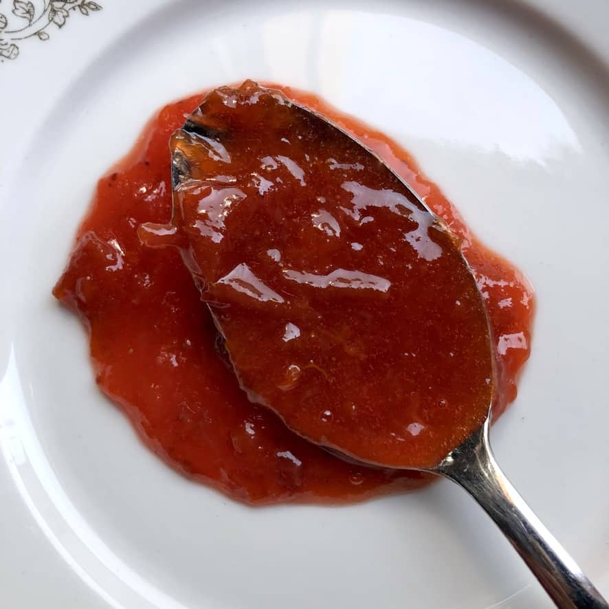 A spoonful of homemade stone fruit and strawberry mixed jam on a spoon on a dessert plate showing the spreadable consistency and the beautiful orange red color of the jam.