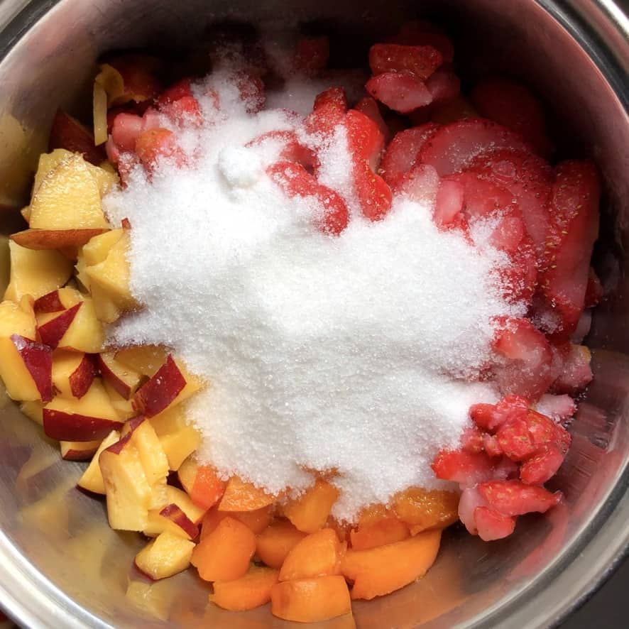 chopped fresh and frozen strawberries, chopped nectarines, and chopped apricots in a pot with sugar poured in the middle.