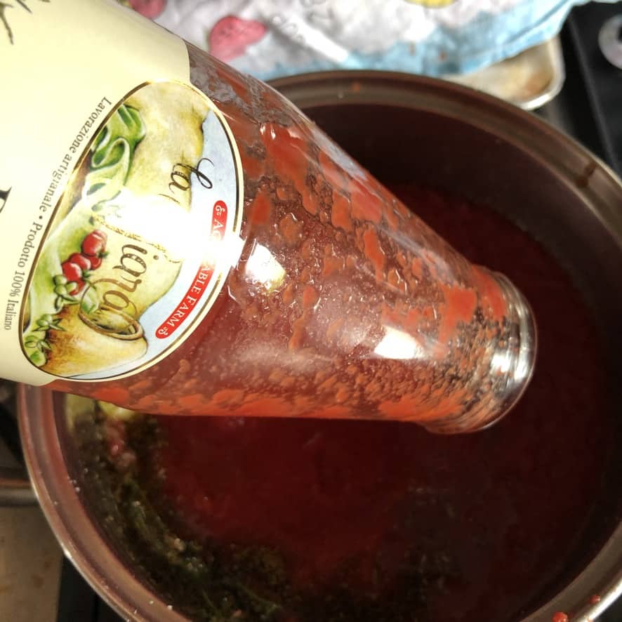 Pouring a jar of organic tomato passata from Abruzzo, Italy into the pot with herbs and olive oil.