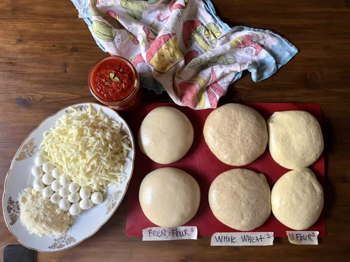 Six total pizza dough balls (00 flour pizza dough, Bread flour pizza dough, and whole wheat pizza dough) lined up on a mat and fully risen ready to be stretched or rolled. Next to the dough is an oval platter filled with mini fresh mozzarella bocconcini balls, freshly grated whole milk mozzarella cheese, and grated Grana Padano and a jar of homemade pizza sauce.