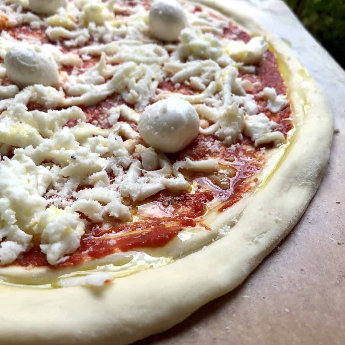 Closeup of thick crust pizza dough made with 00 flour topped with tomato sauce, freshly grated whole milk mozzarella, grana padano, and mini buffalo mozzarella balls before baking.