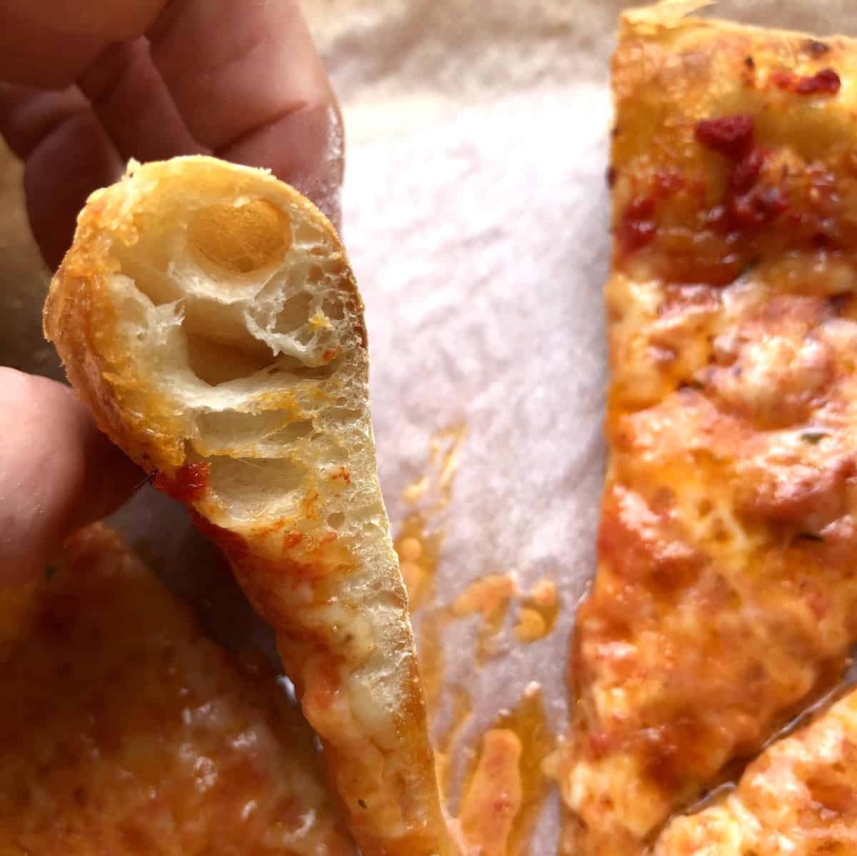 Hand holding a slice of thick crust pizza showing the soft tender interior crumb and fluffy thick crust homemade pizza dough made with 00 flour.
