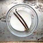 two vanilla beans lying on top of sugar in a glass mixing bowl.