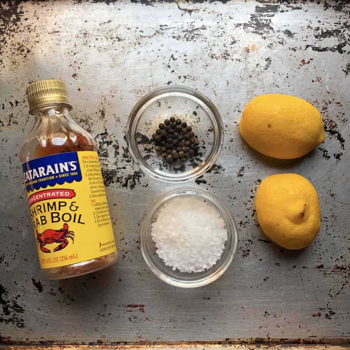 Zatarain's Liquid Crab Boil bottle next 2 small prep bowls with large grain sea salt and black peppercorns, and a halved lemon for seasoning crab for a crab boil.