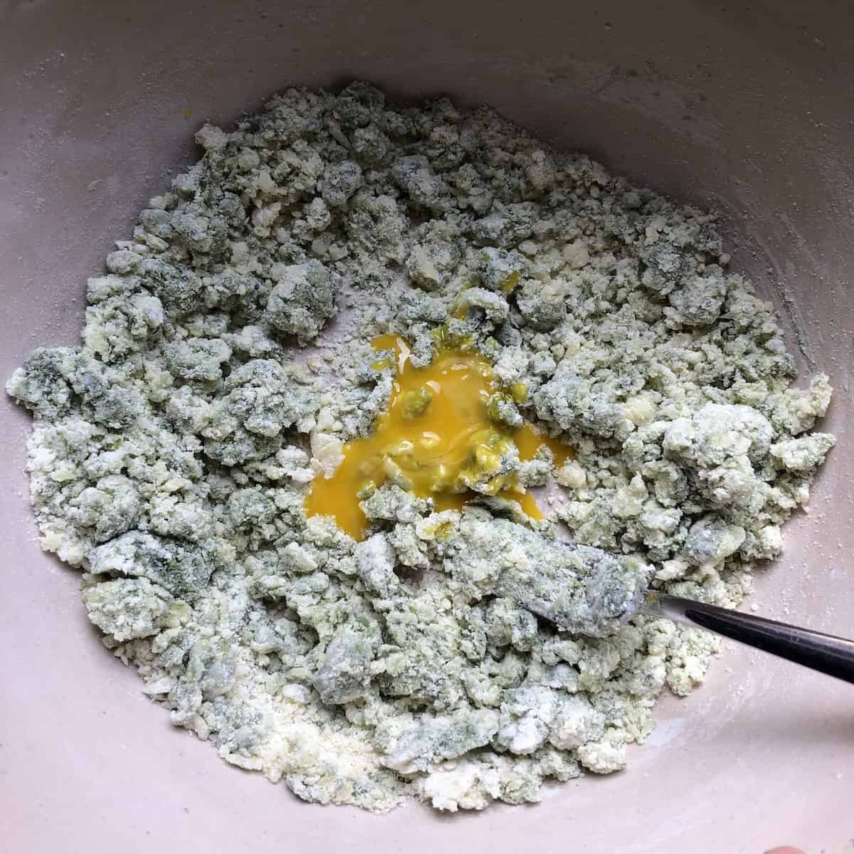
Clumpy green spinach gnocchi mixture in a mixing bowl with an egg yolk added, how to make gnocchi dough from scratch.
