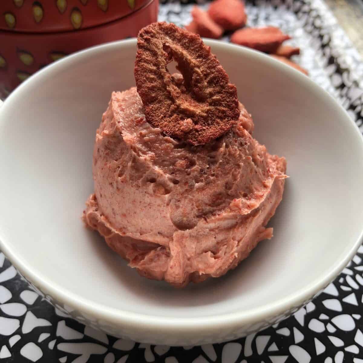 Strawberry butter just whipped and scooped into a small white prep bowl using a portioning scoop with a freeze dried strawberry slice on top for garnish and strawberries in the background with a strawberry tin holder barely visible in the background.