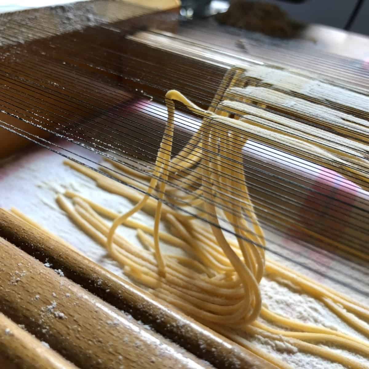 Chitarra spaghetti being cut on my chitarra pasta cutter passed down to me from Luca's parents with square pasta noodles falling through the wires onto the table,showing the sideview of how to make chitarra pasta from scratch.
