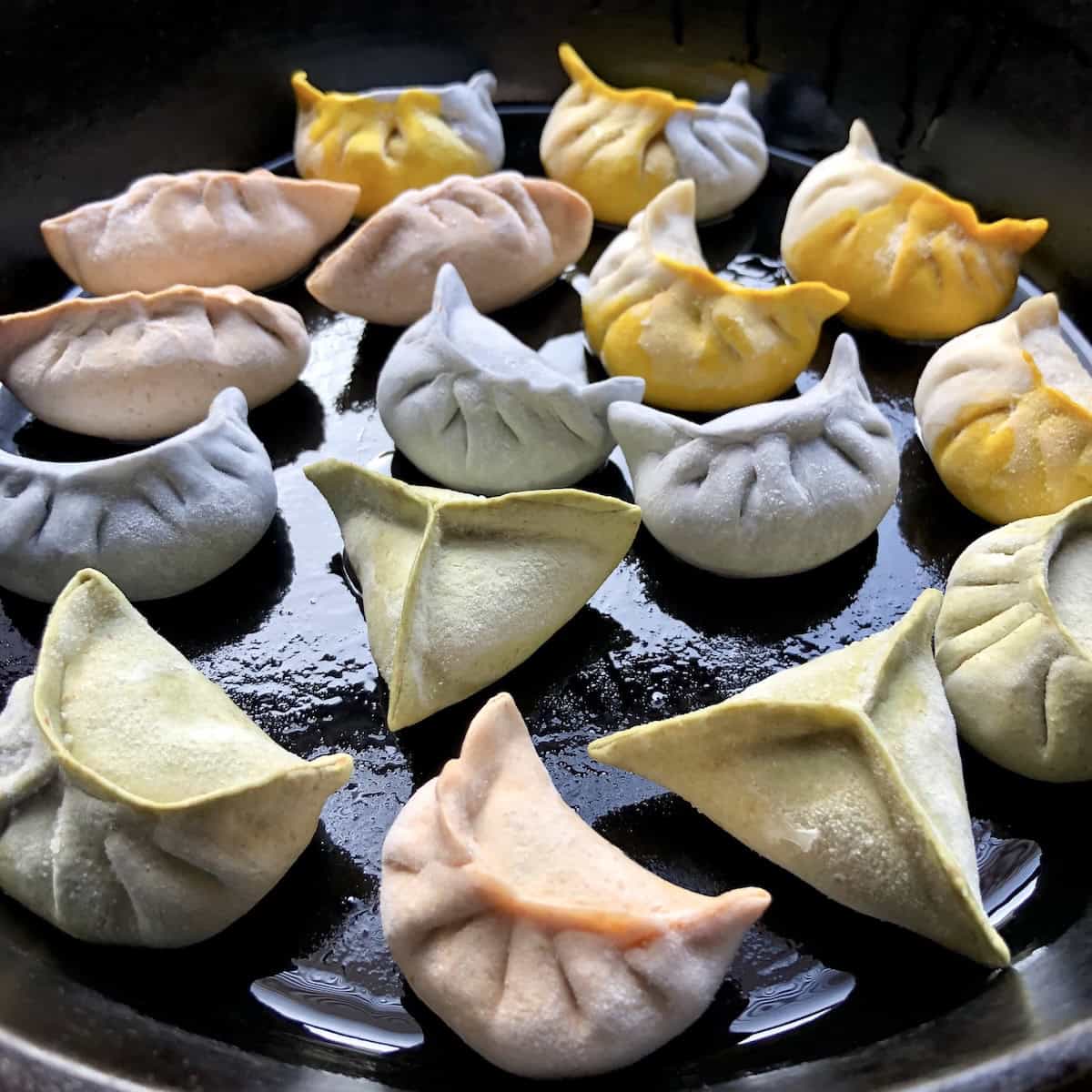 Homemade rozen meal prepped naturally colored dumplings including tie-dyed, gold, pink, purple, green, and white ready to steam as potstickers in a cast iron skillet showing how to make chinese dumplings.
