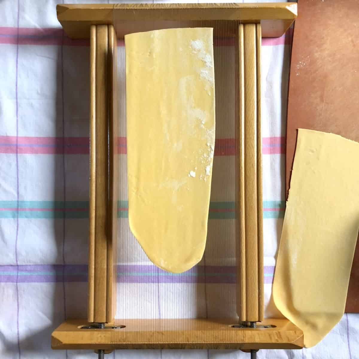 A flat sheet of pasta dough resting on top of the chitarra pasta cutter with another rolled sheet lying next to the guitar ready to cut next, spaghetti alla chitarra recipe.
