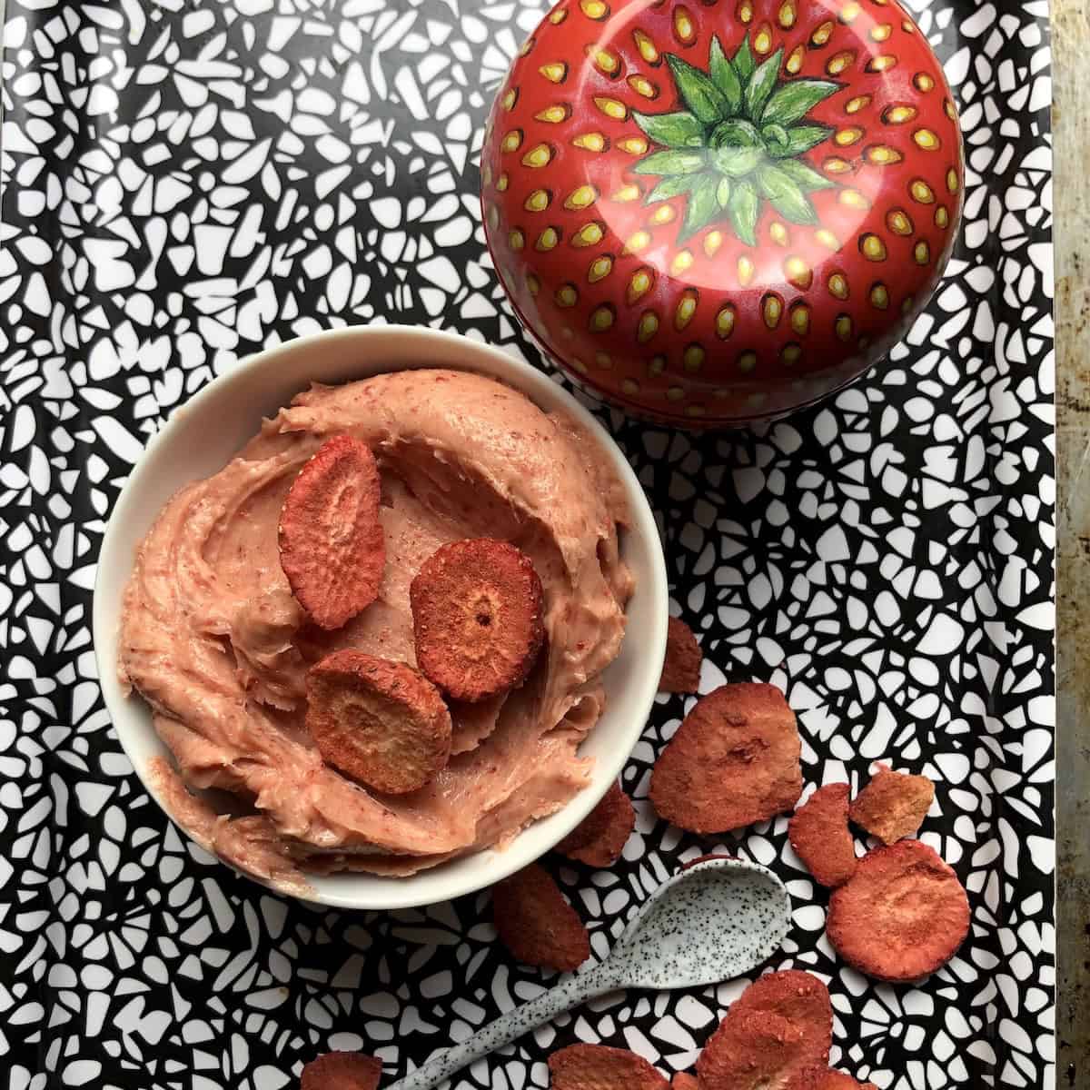 Strawberry butter slathered into a white bowl with freeze dried strawberry slices garnished over the top and a strawberry shaped tin in the background with more freeze dried strawberries scattered with a speckled robin's egg blue serving spoon. 


