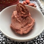 Strawberry butter scooped into a white bowl (looks like dark pink ice cream) with a freeze dried strawberry slice garnish and a strawberry shaped tin in the background with more freeze dried strawberries scattered.