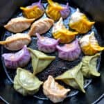 Colored Chinese dumplings being pan-seared and steamed to make potstickers including purple dumplings, green, pink, and two-toned wrappers with half-purple and half gold and half gold and half white dumplings.