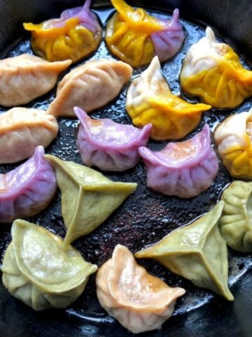 Colored Chinese dumplings being pan-seared and steamed to make potstickers including purple dumplings, green, pink, and two-toned wrappers with half-purple and half gold and half gold and half white dumplings.