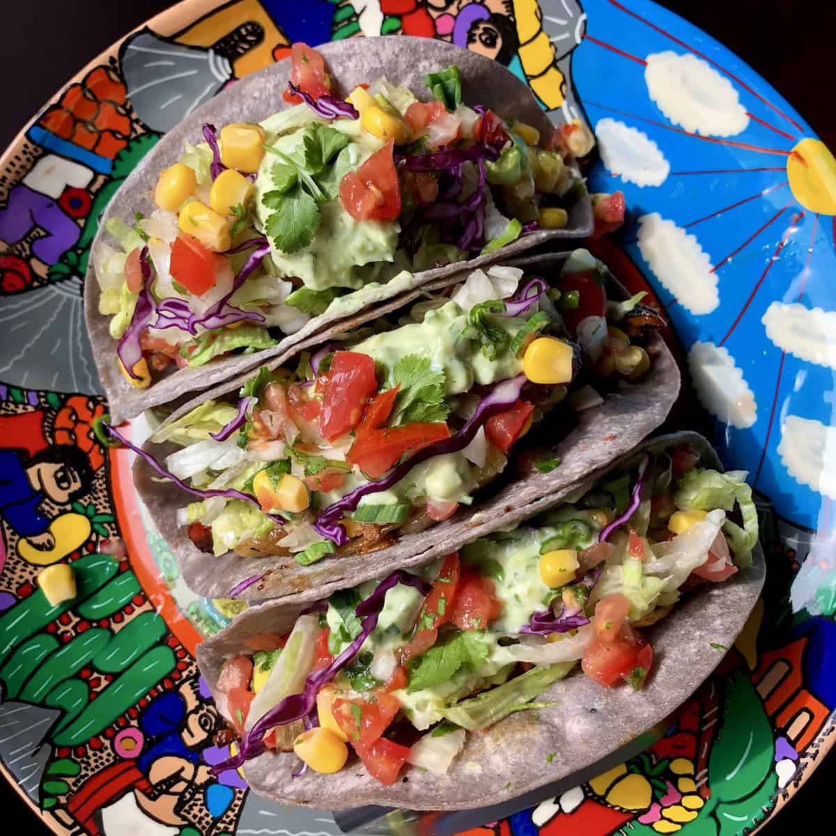 Three vegetarian tacos on a brightly illustrated serving platter with blue corn tortillas.