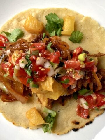A single taco al pastor on a plate consisting of a warm yellow corn tortilla topped with homemade pork pastor, salsa, grilled pineapple, and cilantro.