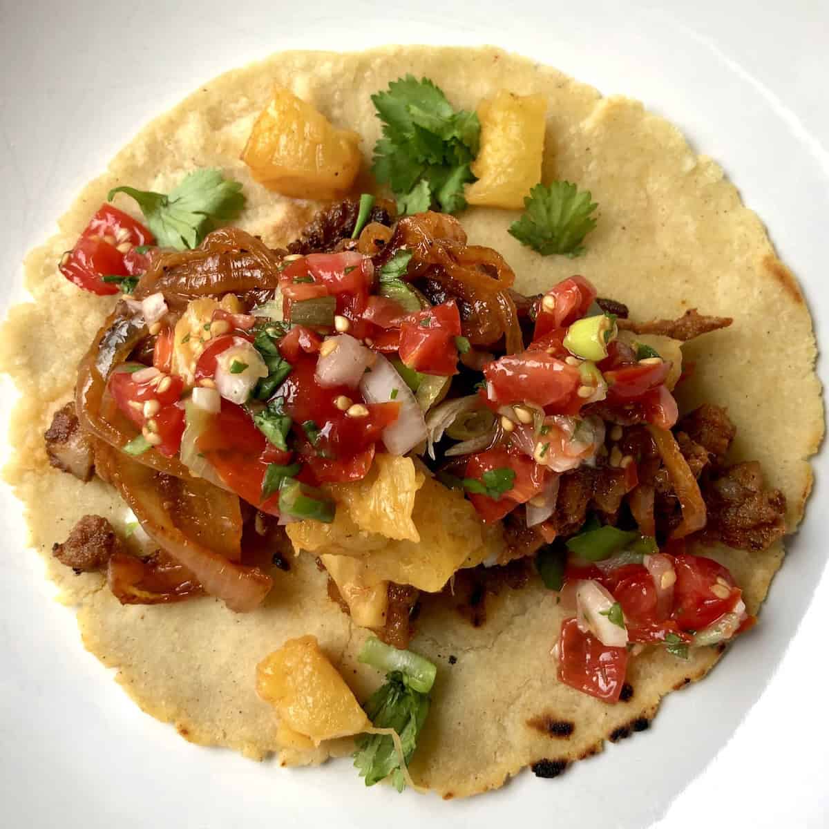 A single taco al pastor on a plate consisting of a warm yellow corn tortilla topped with homemade pork pastor, salsa, grilled pineapple, and cilantro.