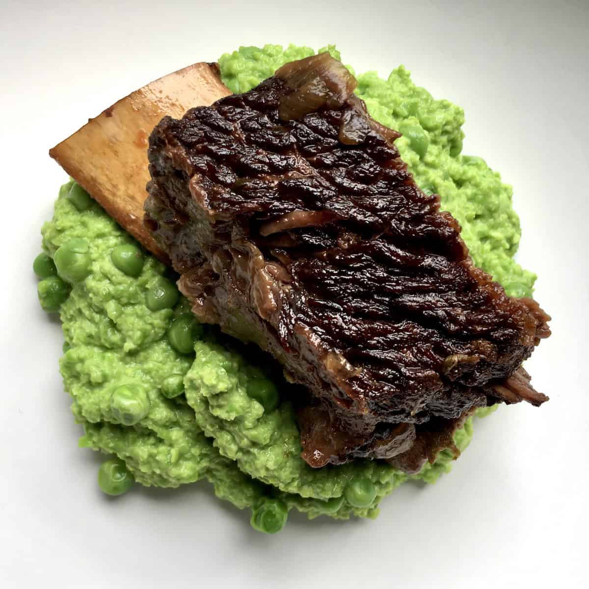 A tender braised beef short rib sitting atop a bed of bright green mushy peas.
