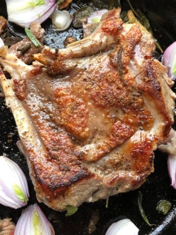 A golden brown spatchcocked turkey leg in a cast iron skillet with olive oil, herbs, and shallots.